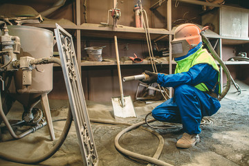 An employee prepares a metal part for painting. A harsh man works in the factory. Sandblast. Blast it.