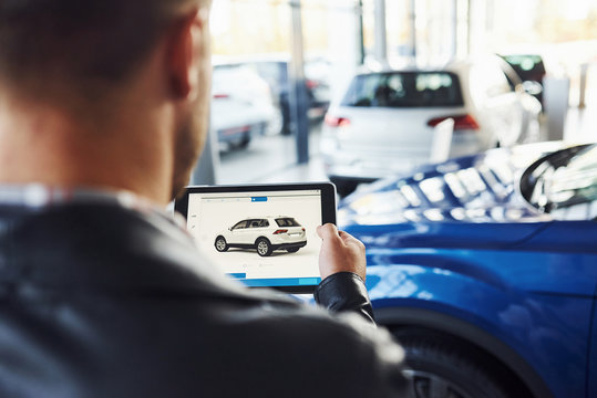 Man stands inside car salon with tablet in hands and looks at the vehicle picture