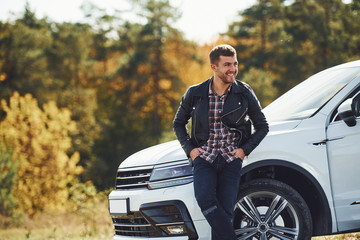 Man in black leather jacket stands near his parked white car outdoors and waits for help