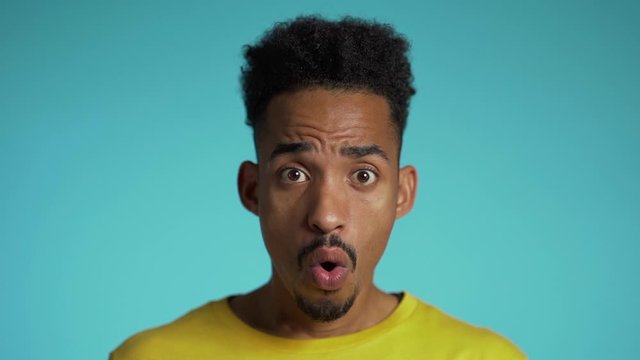 Amazed mixed race man shocked, saying WOW. Handsome african american guy with afro hair surprised to camera over blue background.