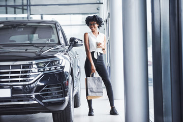 Young african american woman in glasses stands in car salon near vehicle with package in hands