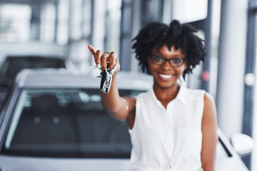 Young african american woman in glasses stands in the car salon near vehicle with keys in hands