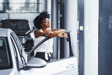 Young african american woman in glasses stands in the car salon near vehicle with keys in hands