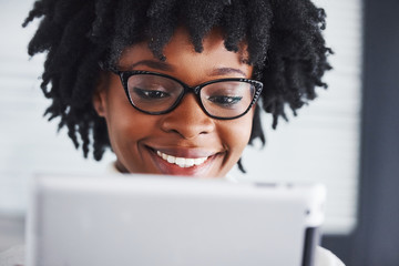 Young african american woman in glasses stands indoors in the office with tablet in hands