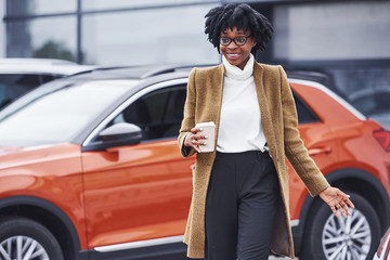 Young african american woman in glasses and with cup of drink stands outdoors near modern car