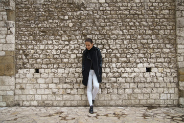 Fototapeta na wymiar Young woman dressed in white jeans and black coat stands leaning on an ancient wall made of stone blocks and looks down