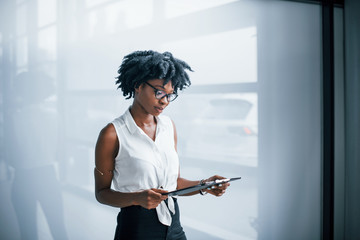 Young african american woman in glasses stands indoors in the office with tablet in hands