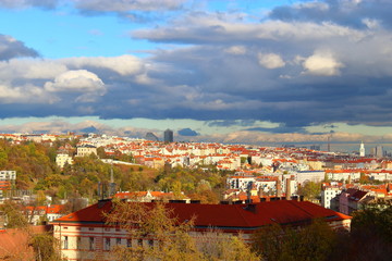 Panorama Of Prague. Houses with orange roofs, under sunlight, among yellowed trees against a blue sky with heavy clouds.