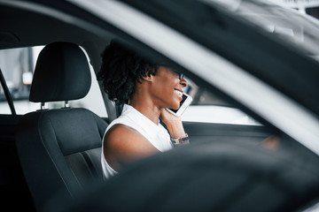 Using phone. Young african american woman sits inside of new modern car