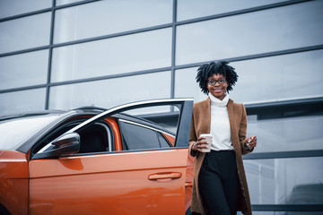Fototapeta na wymiar With cup of drink. Young african american woman in glasses stands outdoors near modern car