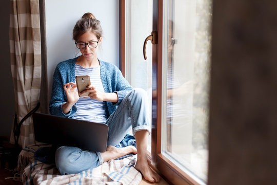 Holiday Shopping Online. Young Woman Makes Purchases In The Internet And Pays By Smartphone On Black Friday. Girl Is Enjoying Buying Gifts And Sitting On Comfortable Windowsill With Laptop At Home.