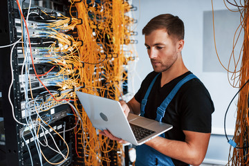 Young man in uniform and with laptop works with internet equipment and wires in server room - obrazy, fototapety, plakaty