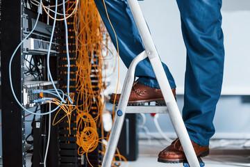 Man in uniform stands on the ladder and works with internet equipment and wires in server room