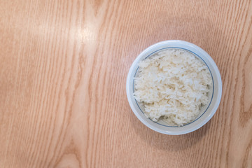 Top view image of white rice in a white bowl with wooden table background