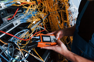 Young man in uniform with measuring device works with internet equipment and wires in server room