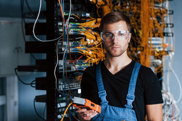 Young man in protective glasses works with internet equipment and wires in server room