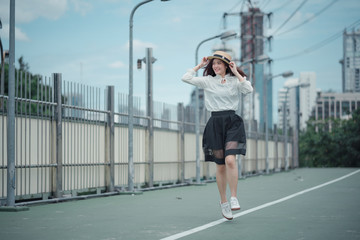 asian girl with long wavy brown hair in white shirt black skirt and boater hat running happily on the green pathway with hands on hat feeling fun on sunny day beautiful blue sky with clouds in summer