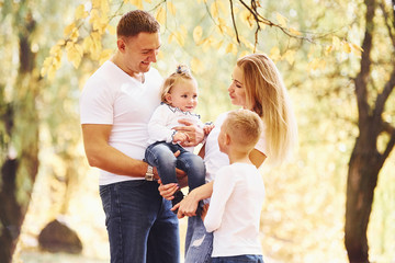 Mother and dad holds kids at shoulders and in hands. Cheerful young family have a walk in an autumn park together