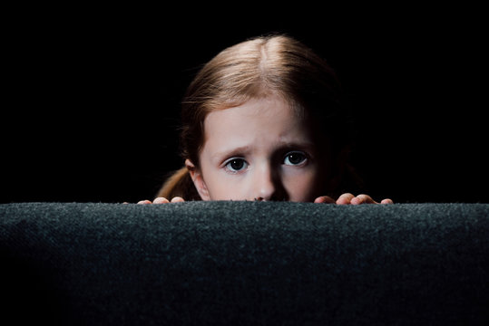 scared child hiding behind armchair and looking at camera isolated on black