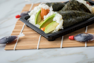 Lachs und Surimi Avocado Temaki Sushi in Lieferdienst Verpackung mit Soja Soße Fische und Sushimatte auf Marmor Hintergrund