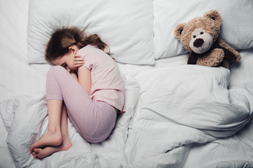 top view of scared child lying on white bedding near teddy bear