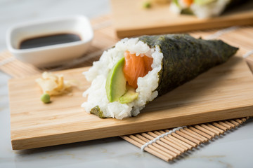 Lachs Fisch Avocado Temaki Sushi, eingelegter Ingwer mit Soja Soße und Sushimatte auf Bambus Brett Teller und Marmor Hintergrund