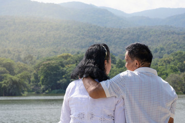 Happy retirement elder couple hugging standing talking together outdoor lake with green mountain nature over sky background.