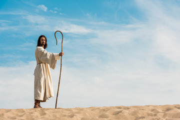 handsome man in jesus robe holding wooden cane against sky in desert