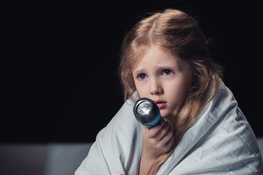 Scared Kid Sitting Under Blanket And Holding Flashlight Isolated On Black