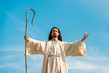 jesus with outstretched hands holding wooden cane against blue sky