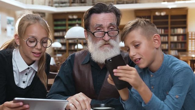 Attractive 12s grandchildren trying to convince respected grandfather about the advantages of modern divices over the books,sitting in the library