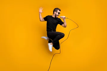 Zelfklevend Fotobehang Full body profile photo of crazy hipster guy jumping high holding microphone music lover singing favorite song wear sun specs black t-shirt pants isolated yellow color background © deagreez