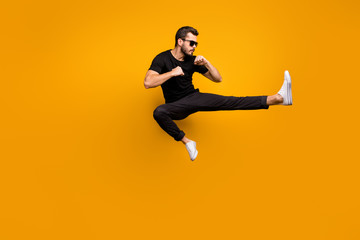 Fototapeta na wymiar Full length photo of handsome guy jumping high practicing self defense kicking confident facial expression wear sun specs black t-shirt pants isolated yellow color background