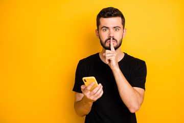 Censorship no telling secret concept. Serious guy blogger hold use cellphone search private fake news show mute quiet sign index finger wear black t-shirt isolated yellow color background - obrazy, fototapety, plakaty