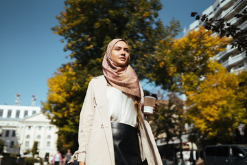 Fototapeta premium a Muslim woman with a smile of hope and joy holds coffee in her hand and goes towards life along the street