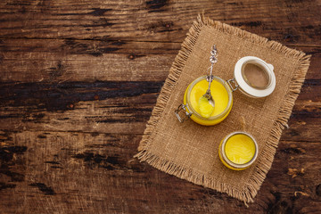Pure or desi ghee (ghi), clarified melted butter. Healthy fats bulletproof diet concept or paleo style plan. Glass jar, silver spoon on vintage sackcloth. Wooden boards background