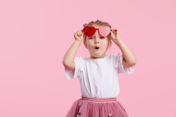Portrait of surprised cute little toddler girl in the heart shape sunglasses. Child with open mouth having fun isolated over pink background. Looking at camera. Wow funny face