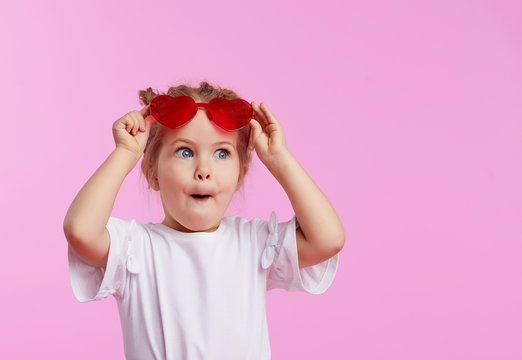 Naklejki Portrait of surprised cute little toddler girl in the heart shape sunglasses. Child with open mouth having fun isolated over pink background. Looking at camera. Wow funny face