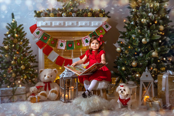 New Year's greetings. A little girl in a red dress and with a red bow in her hair sits near a Christmas tree and fireplace and reads a book.