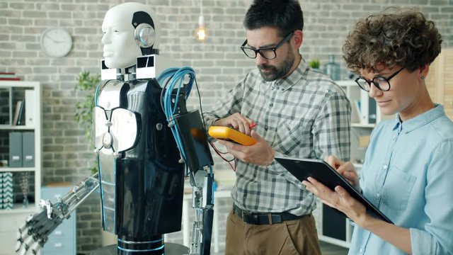 Scientists man and woman are checking robot with electronic devices using tablet in office room concentrated on research work. People and technology concept.