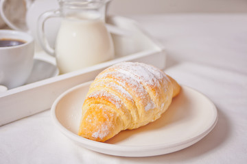 Fresh croissant bun on the white plate with cup of coffee, jar of milk on the wooden tray on the background.