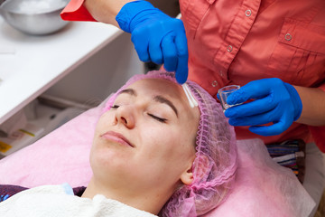 A young girl is lying on a couch during cosmetic procedures with a mask on the face above which beautician woman applies acid peeling. Cleansing and make up.