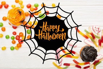 top view of colorful gummy sweets, cupcakes and bonbons on white wooden table with spiderweb and happy Halloween illustration