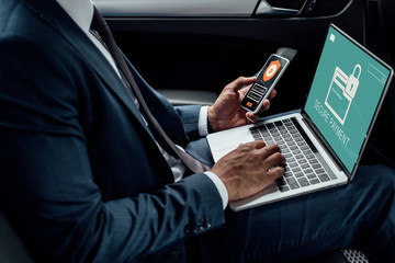 cropped view of african american businessman using laptop and smartphone in car with cyber security illustration