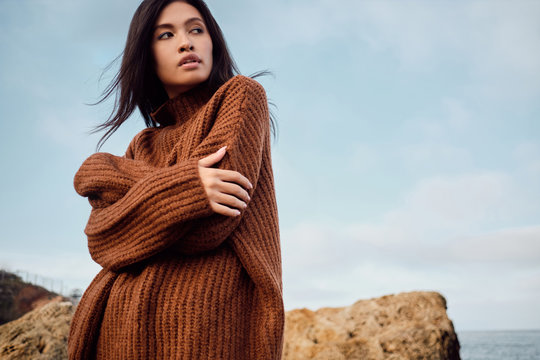 Beautiful casual Asian girl in knitted sweater dreamily looking away by the sea