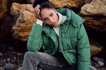 Obraz premium Portrait of casual African American girl in stylish down jacket intently looking in camera while posing on stones by the sea
