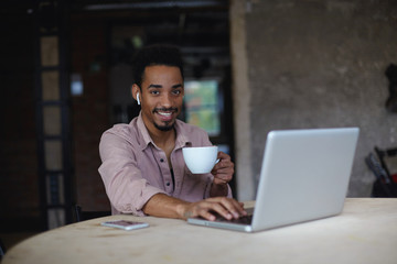 Photo of cheerful pretty young bearded male with dark skin working remotely with laptop in coworking space, being in nice mood and smiling sincerely to camera with cup of tea in his hand