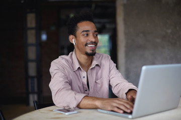 Photo of charming young dark skinned male with short haircut sitting at table and keeping hands on...