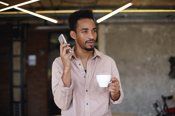 Photo of attractive dark skinned bearded guy with short haircut keeping cup of coffee in raised hand, looking thoughtfully ahead and going to make call with his smartphone