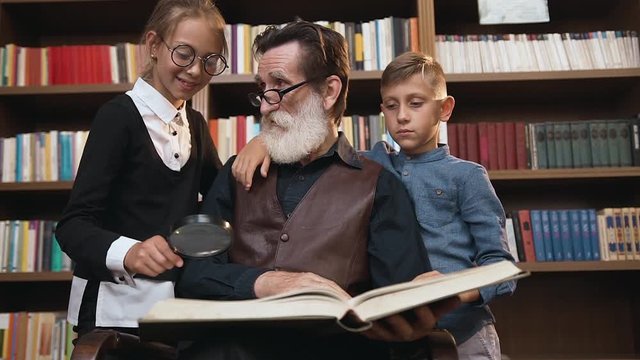 Lovely teenage schoolchildren reading book with their intelligent old grandfather near library bookshelves